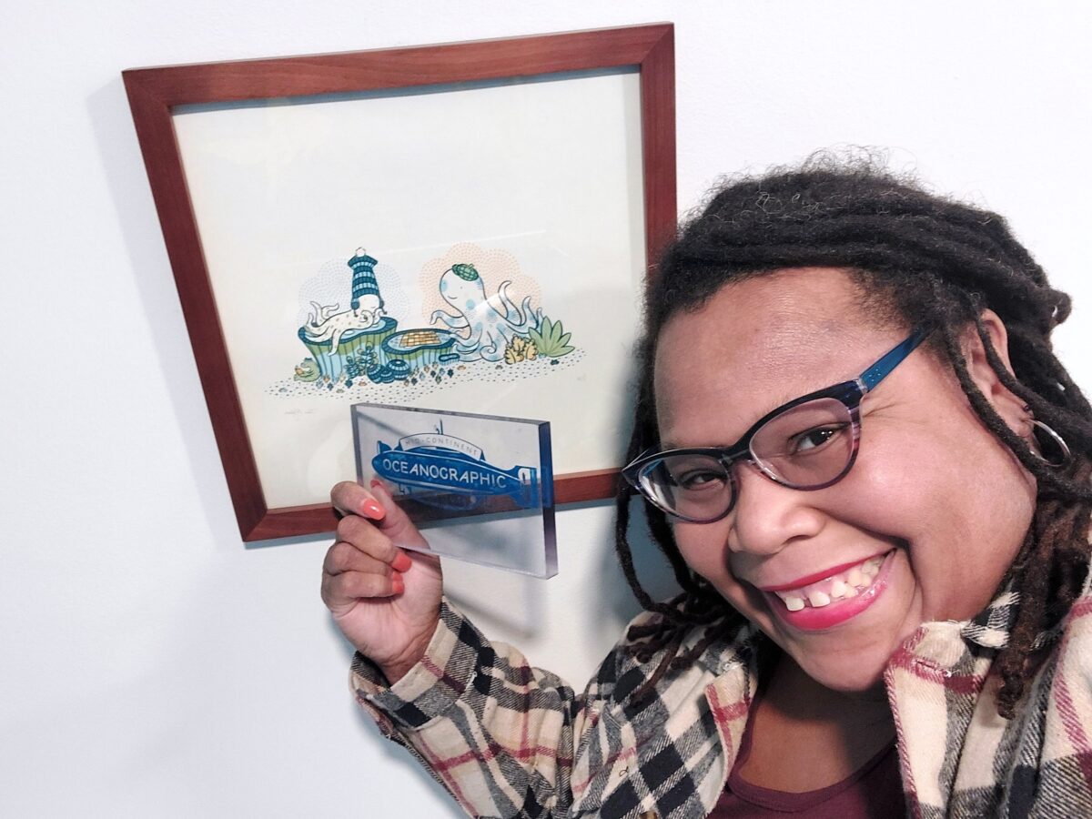 Smiling Black woman in glasses holding up an "826 MSP" postcard in front of a framed illustration
